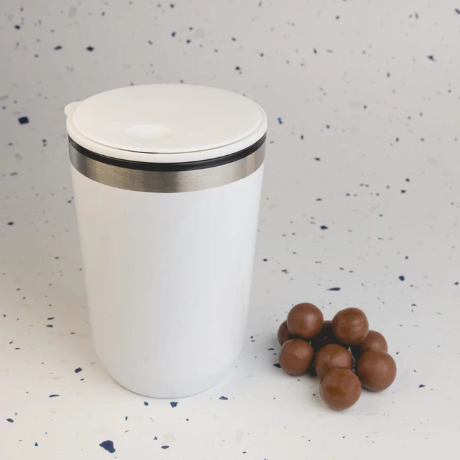 A reusable white coffee cup with a stainless steel band, beside a small pile of brown chocolate balls.