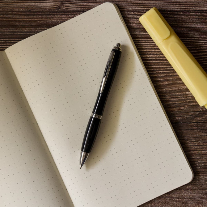 A black plastic pen lies on a blank notebook alongside a yellow highlighter.