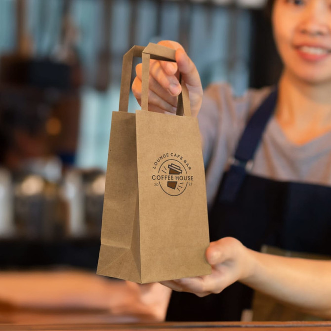 A small, brown paper bag with flat handles and a logo, being held by a person in an apron.