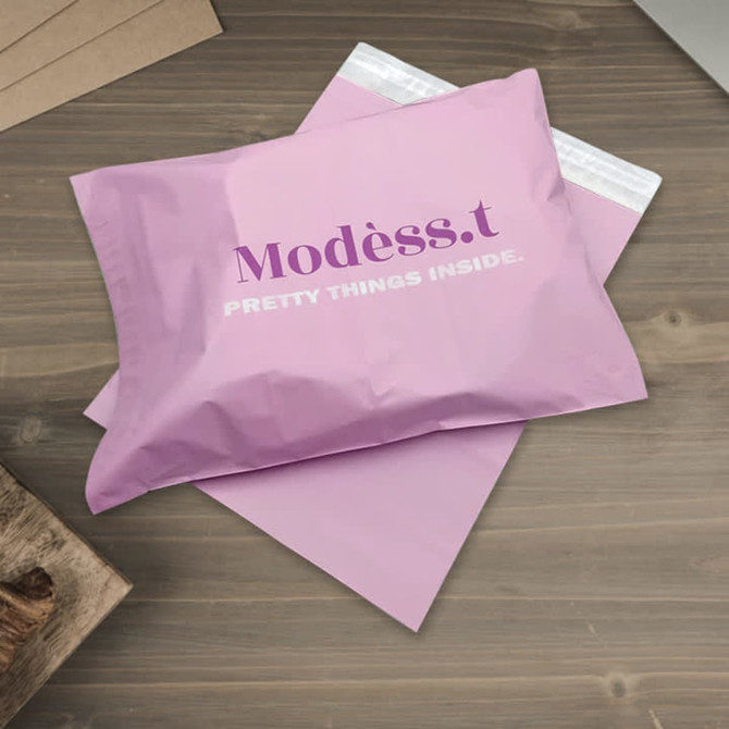 A large biodegradable mailer bag in pink with a logo, resting on a wooden surface with a brown background.