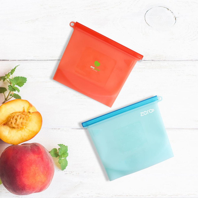 Two silicone reusable storage pouches in red and blue, placed on a wooden surface alongside a peach and a sprig of herbs.