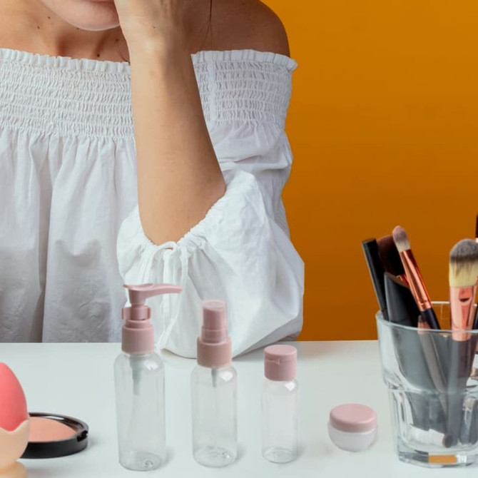 A cosmetic travel bottle kit featuring clear bottles with pink lids, alongside makeup brushes and tools on a bright yellow background.