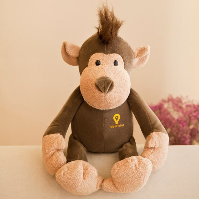 A brown and beige monkey plush toy with a tuft of hair and a logo on its chest, sitting on a surface.