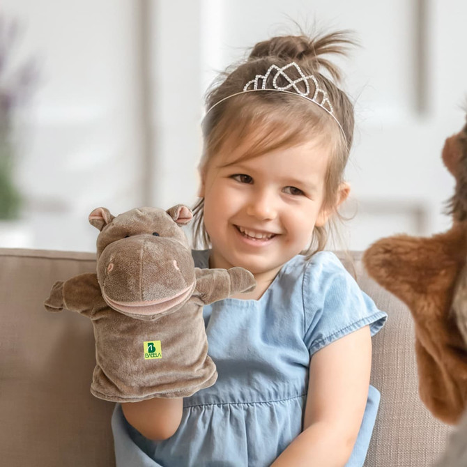 A plush hippopotamus hand puppet in brown, held by a smiling child wearing a blue dress, with a decorative headband.