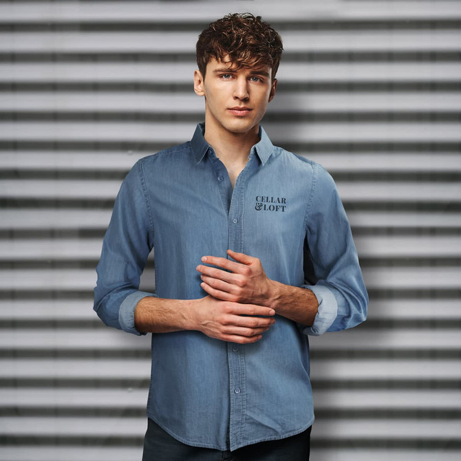 A denim button-up shirt in light blue, featuring a logo on the chest, modelled by a man against a striped background.