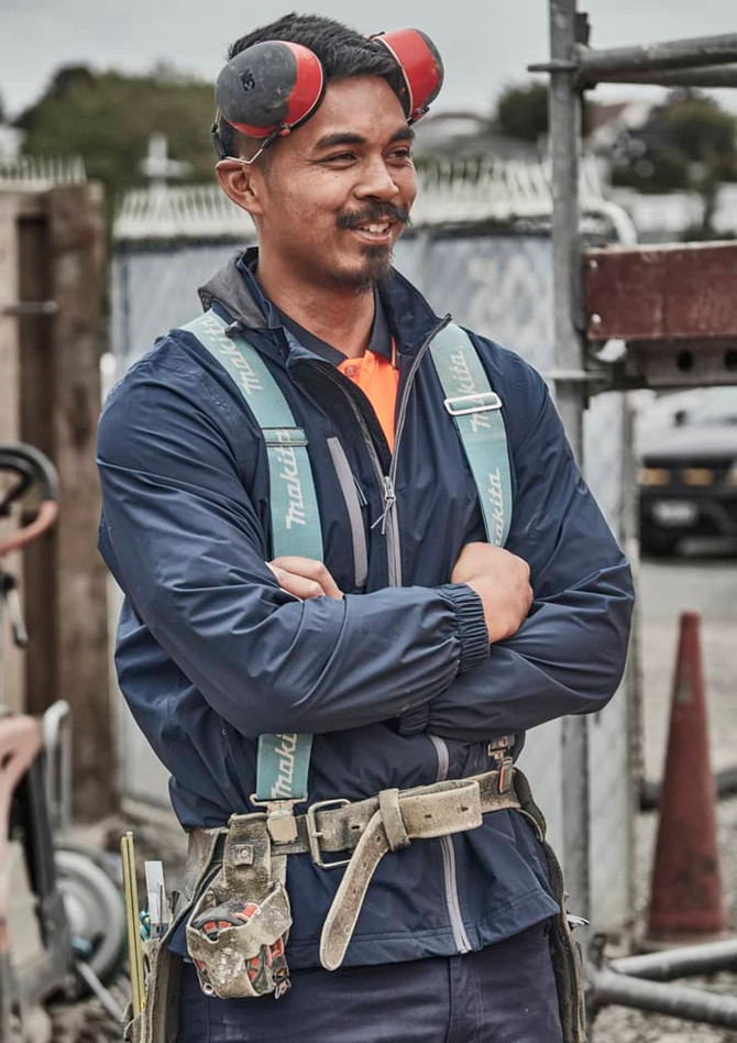 A man in a navy blue packable rain jacket stands with arms crossed, wearing earmuffs and a safety harness.