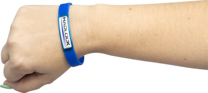 A blue silicon wristband on a wrist, featuring a logo on an attached rectangular plaque.