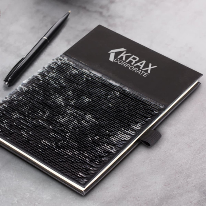 A sequin notebook in black with a logo, accompanied by a black pen on a textured grey surface.