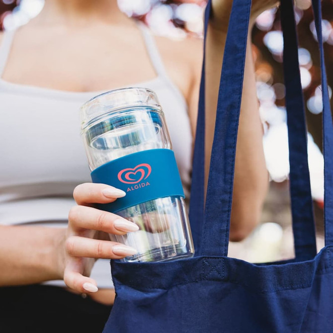 A reusable coffee cup with a transparent body and a blue silicone sleeve, held within a navy tote bag.