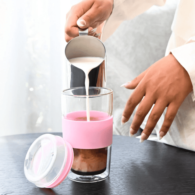 A reusable coffee cup in clear glass with a pink silicone sleeve, being filled with milk.