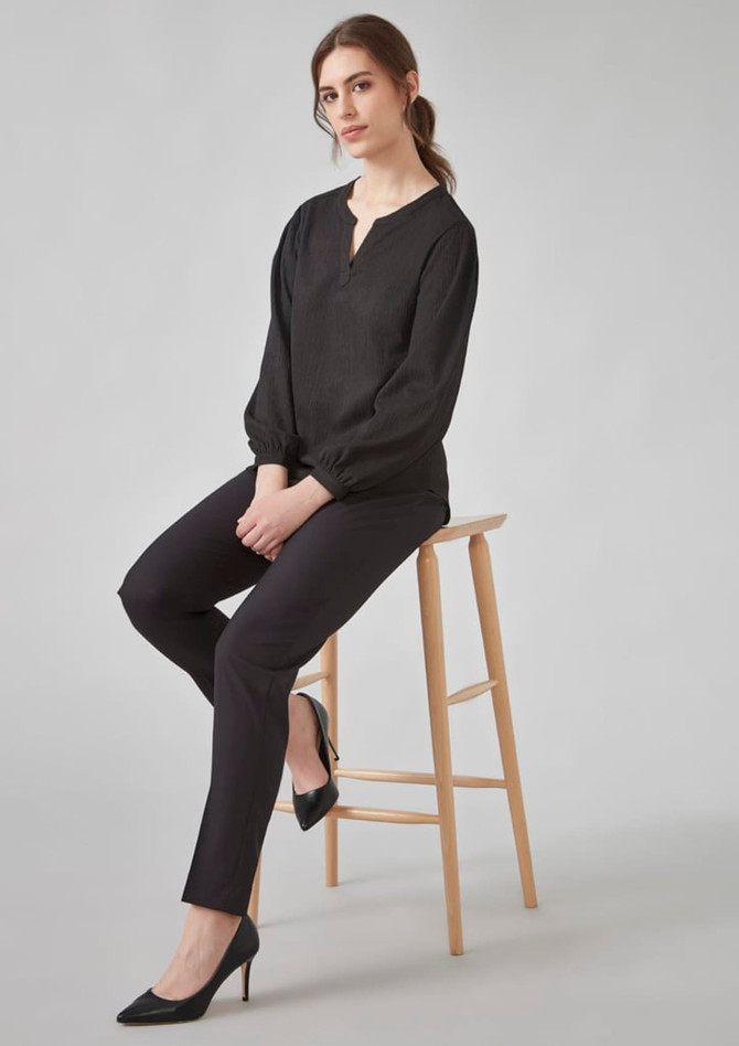 A black long sleeve blouse with a ruched design, paired with slim black trousers and heels, seated on a wooden stool.