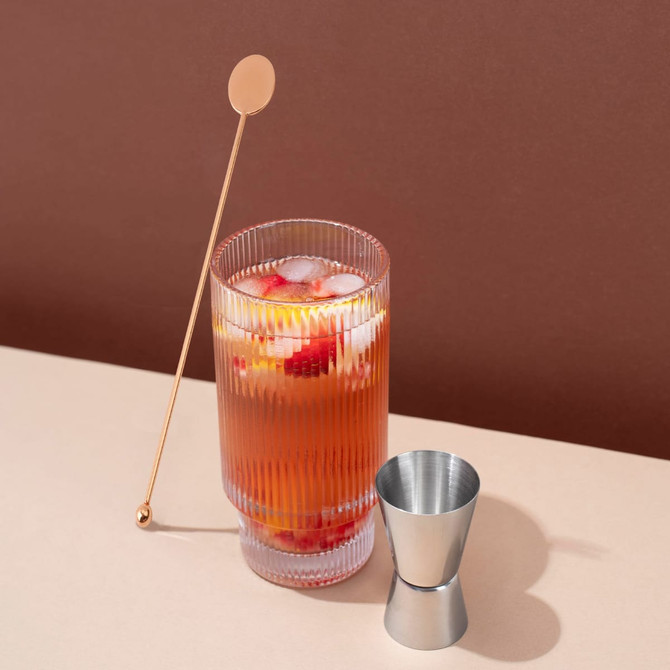 A tall, ribbed glass filled with a colourful cocktail, accompanied by a stainless steel cocktail stirrer and jigger.