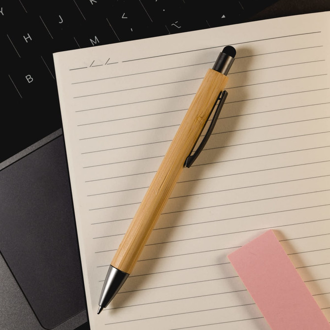 A bamboo pen with a black stylus tip, placed on a lined notepad next to a pink sticky note and a laptop.
