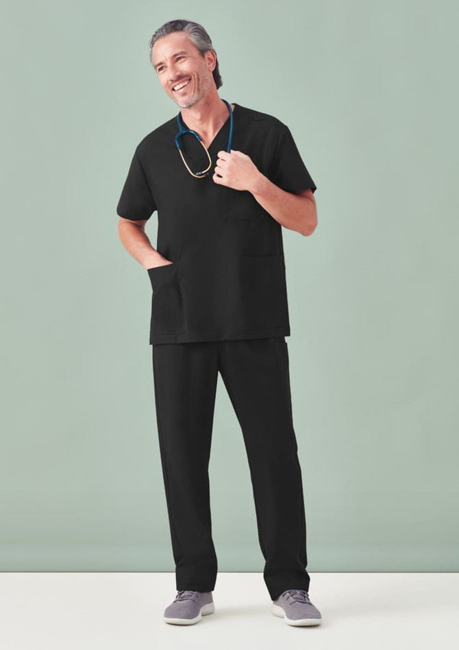 A man wearing black unisex scrub pants paired with a black scrub top, standing against a light green background.