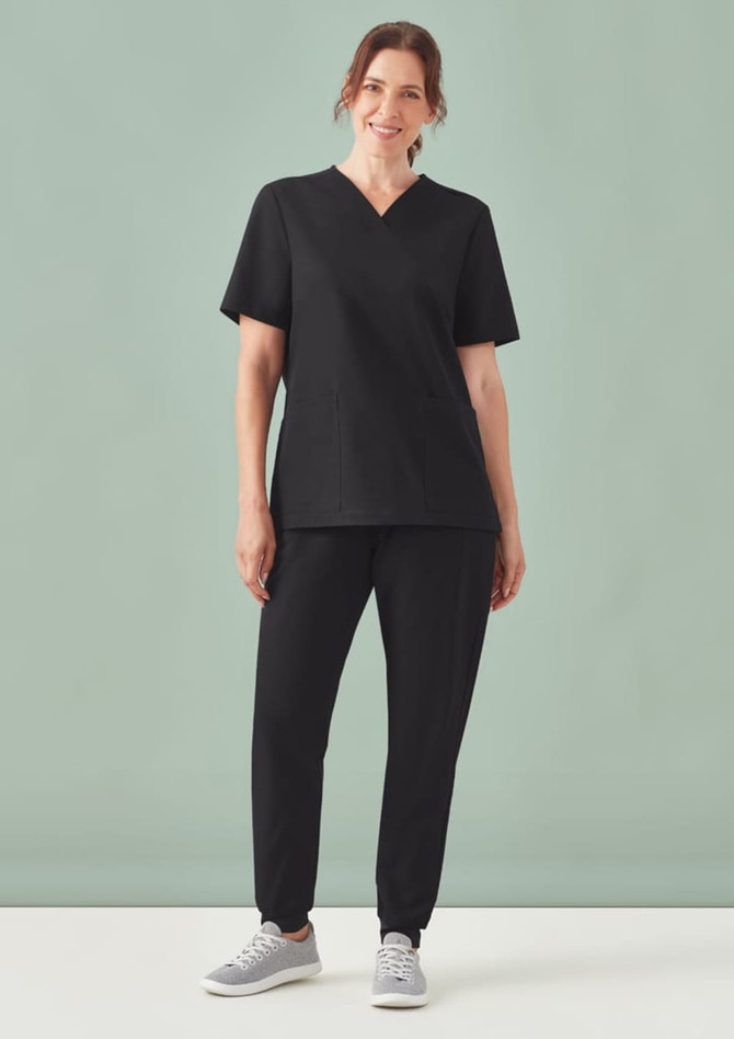 A woman wearing black jogger scrub pants and a matching top, standing against a pale green background.