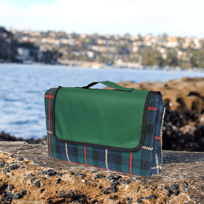 A green picnic rug, featuring a tartan plaid pattern in blue, red, and cream, rests on a rocky surface by water.