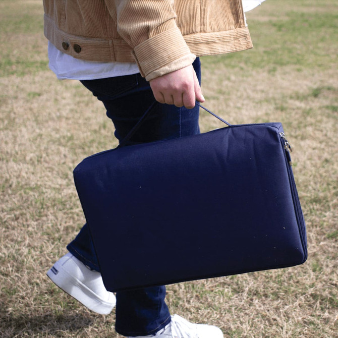 A person carries a navy blue picnic rug with a waterproof backing, featuring a handle for easy transport.