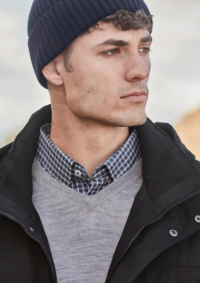 A man wearing a navy beanie, grey sweater, and a checked shirt. The background features a blurred outdoor setting.