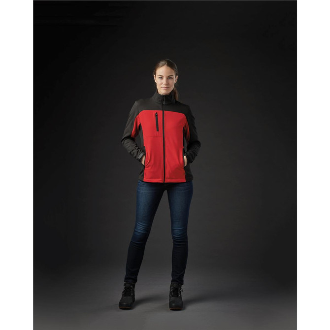 A woman wearing a red and black softshell jacket with pockets, standing against a dark background.