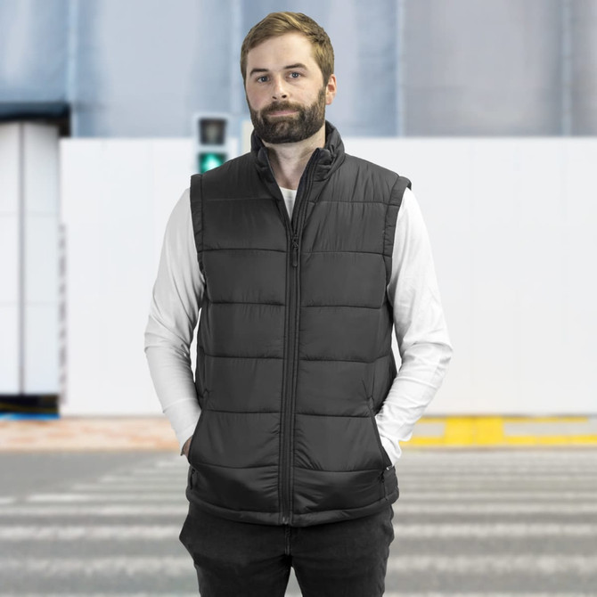 A man wearing a black puffer vest stands against a neutral background. The vest features a zip front and a logo.