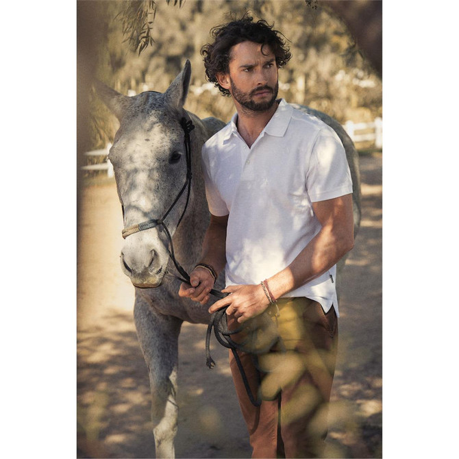 A man wearing a white cotton polo shirt stands beside a grey horse, set in a natural outdoor environment.