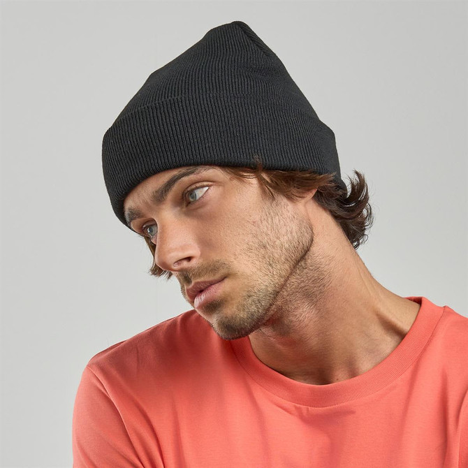 A black ribbed beanie is displayed on a model wearing a coral top, with a focused expression.