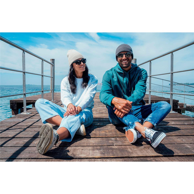 A couple sits on a pier wearing light grey and dark grey beanies, with casual clothing and sunglasses, by the water.