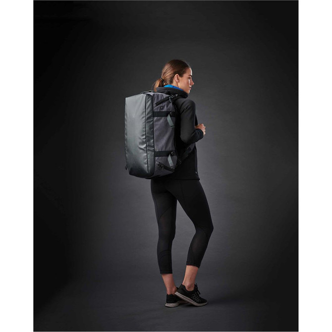 A woman wearing black activewear carries a grey Equinox 30 Duffel Bag on her back against a dark background.