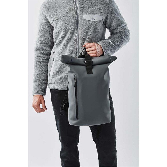 A person holding a grey Sargasso backpack with a roll-top design and a front zip pocket.