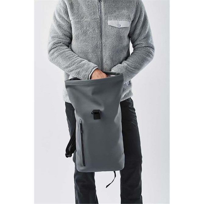 A grey Sargasso backpack held by a person wearing a fleece jacket, featuring a side zip pocket and adjustable strap.