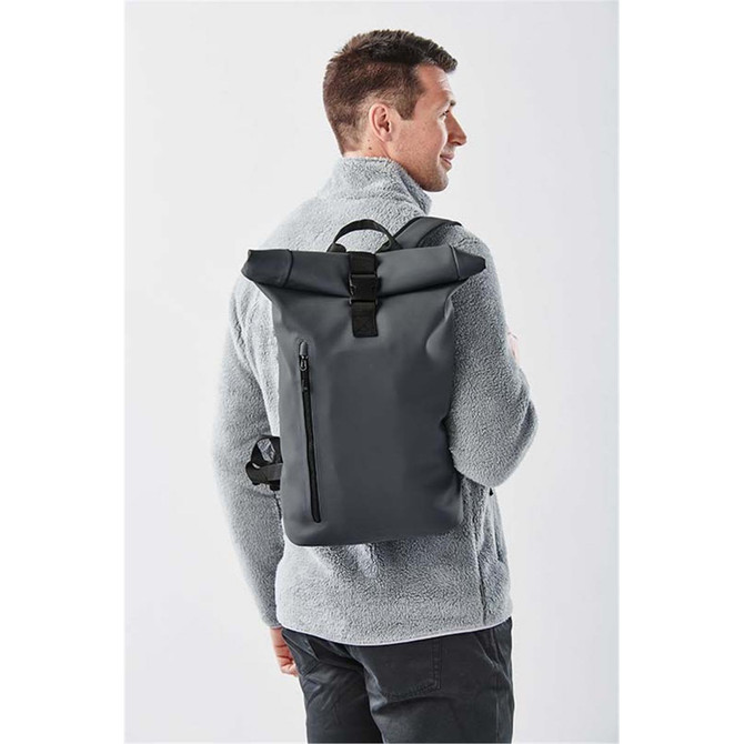 A grey roll-top backpack is worn by a man, showing a zippered front pocket and featuring a logo.