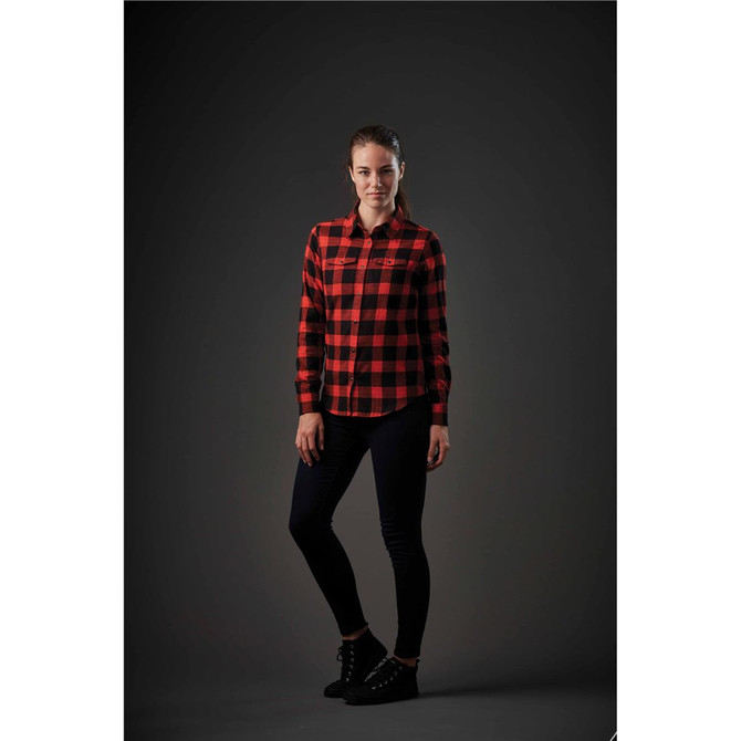 A model in a red and black checkered button-up shirt poses against a dark background, paired with dark jeans and ankle boots.