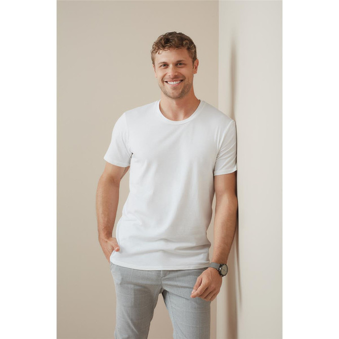A man in a white crew neck t-shirt leans against a wall, smiling and wearing light grey pants.