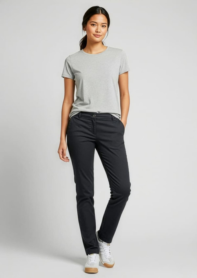 A woman stands in black chino pants and a grey t-shirt, smiling with one hand in her pocket. The image includes a logo.