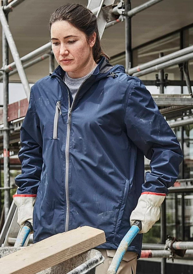A woman in a navy blue packable rain jacket, wearing gloves, stands near scaffolding, holding a wooden beam.