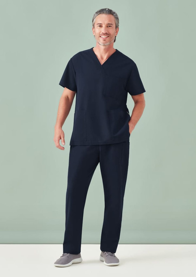 A man wearing navy unisex scrub pants and a matching short-sleeve top, standing against a light green background.