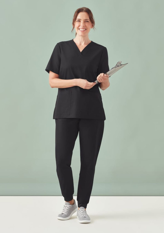 A woman wearing black women's Pulse jogger scrub pants and a matching top, holding a clipboard, standing against a light green background.