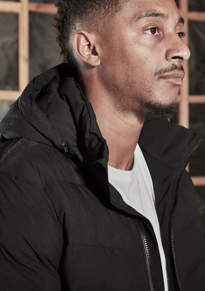 A black hooded puffer jacket, featuring a zipper and a discreet logo, worn by a model.