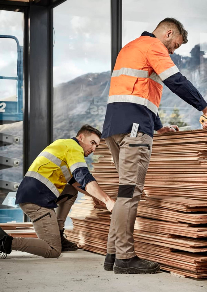 Two men in workwear, one in a yellow and navy shirt and the other in an orange and navy shirt, examine wooden panels while wearing beige stretch pants.