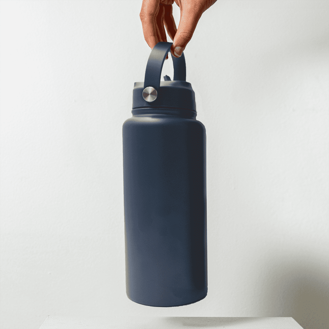 A drink bottle in dark blue, featuring a handle and a silver accent, held by a hand against a plain background.