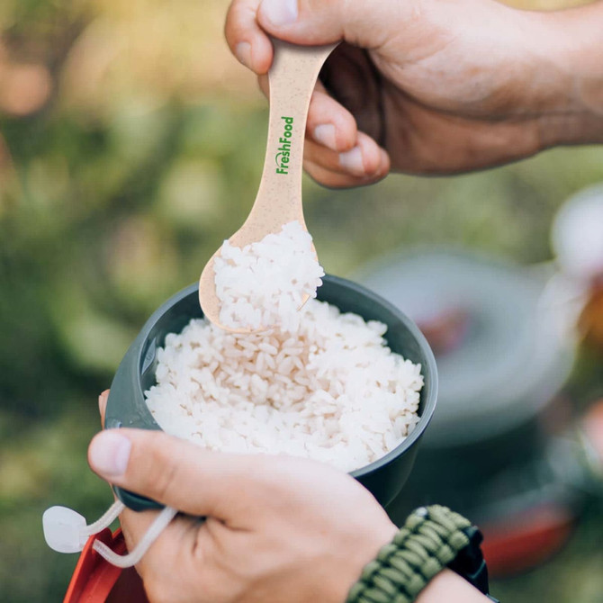 A hand holds a beige spoon scooping rice from a gray bowl outdoors. The spoon features a logo.