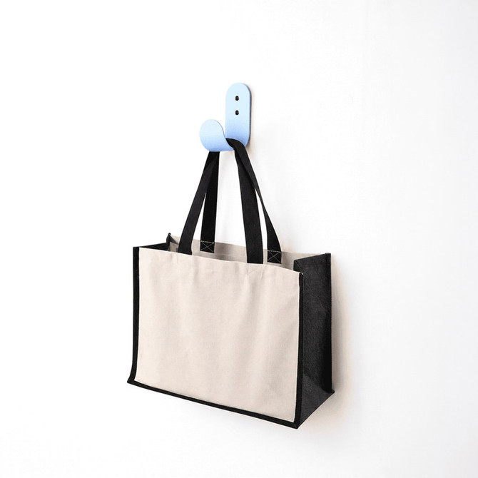 A functional tote bag in beige and black hangs on a blue hook against a white wall. The bag has handles and a logo.