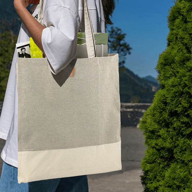 A beige and cream tote bag with long straps, featuring a logo, held by a person outdoors.