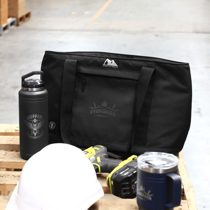 A black cooler bag and drink bottle sit on a wooden pallet, accompanied by tools and a white cap. The bag has a logo.