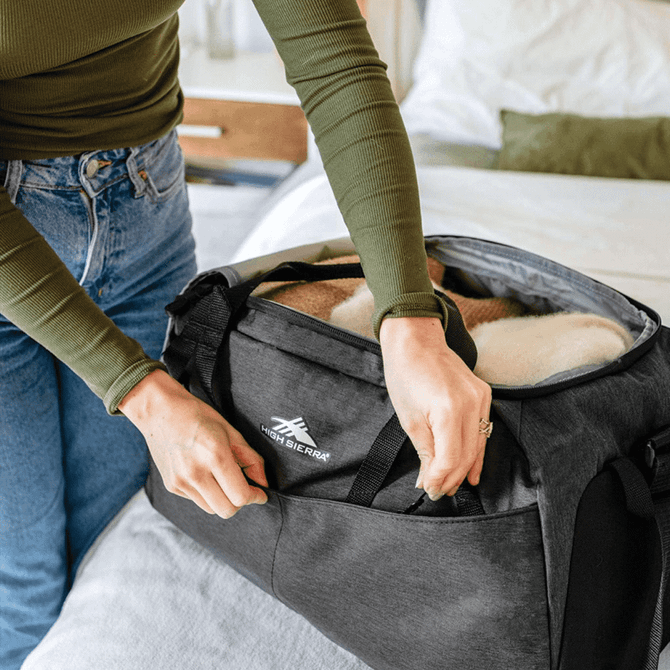 A person opens a charcoal grey duffel bag, revealing a cozy interior with a blanket inside. The bag has a logo.