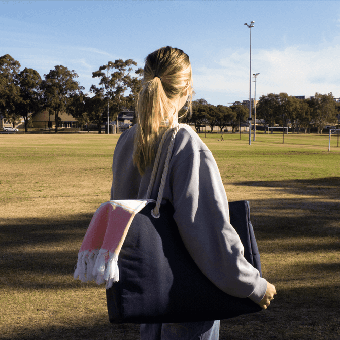 A person holds a navy blue beach bag with a pink and white tassel, standing in a park on grass.