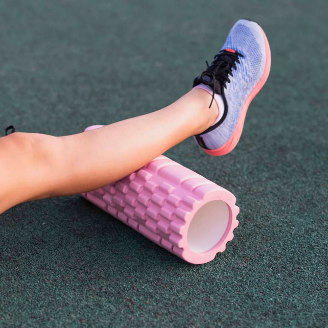 A pink foam yoga roller positioned on green flooring, with a person's leg resting on it, wearing blue running shoes.