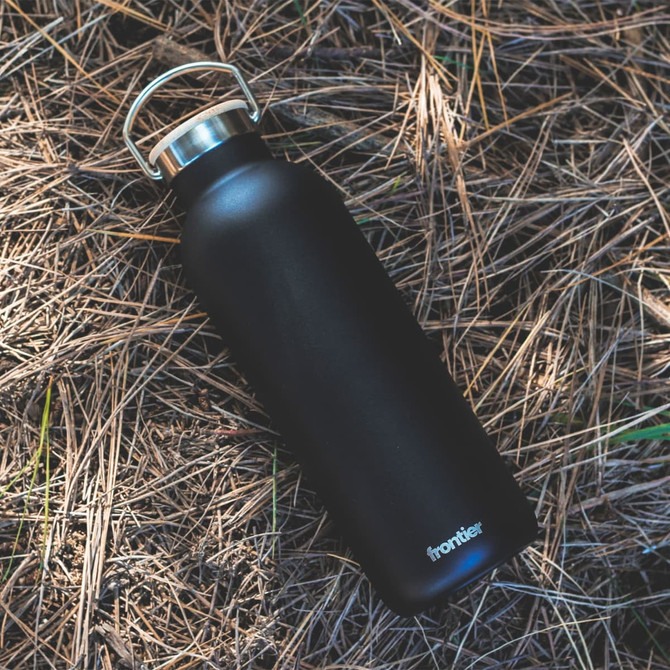 A drink bottle in matte black with a silver cap, featuring a logo. Set on a bed of pine needles.