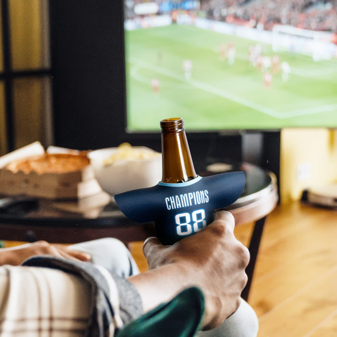 A drink bottle is in a navy blue Jersey Can Cooler with a logo, set next to a TV displaying a sports event.