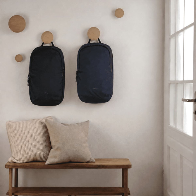 Two dark blue backpacks hang on a wall, alongside wooden hooks, with a bench and cushions in the foreground.